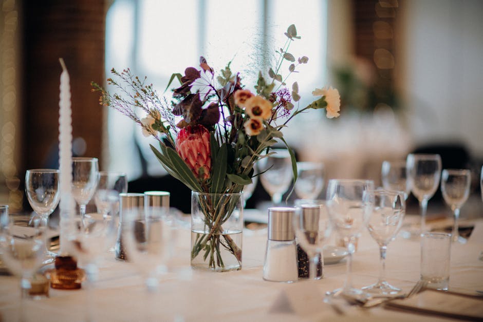 A floral arrangement featuring a variety of fresh flowers in a clear glass vase, placed on a table set for a special event. The bouquet includes a large pink protea, purple and white wildflowers, and 